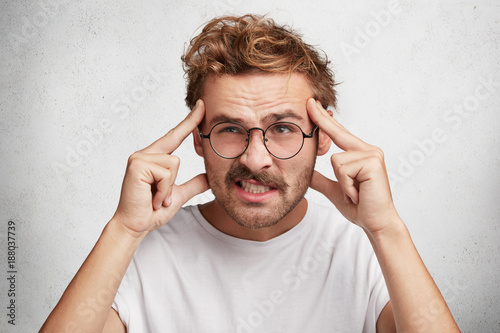 Bild auf Leinwand Handsome male tries to concentrate and remember some details, keeps finger on temples and clenches teeth, frowns face with dissatisfaction as being forgetful and absent minded