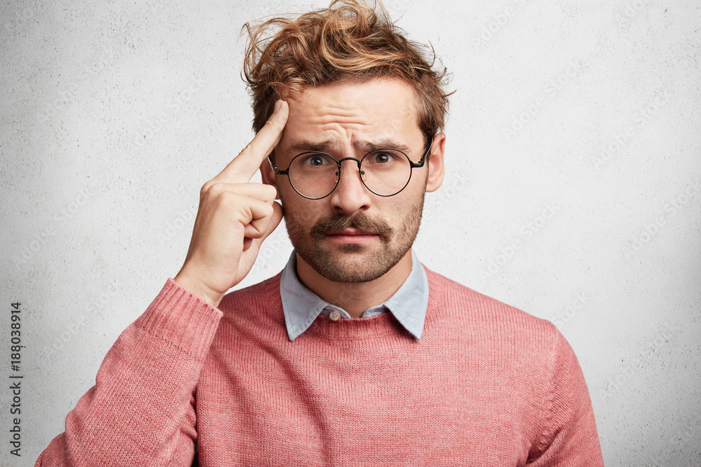 Portrait of handsome young stylish male tries to concentrate as sees something in front, has problematic expression, dressed in pink sweater, has round eyewear, isolated over white concrete wall Portrait of handsome young stylish male tries to concentrate as sees something in front, has problematic expression, dressed in pink sweater, has round eyewear, isolated over white concrete wall