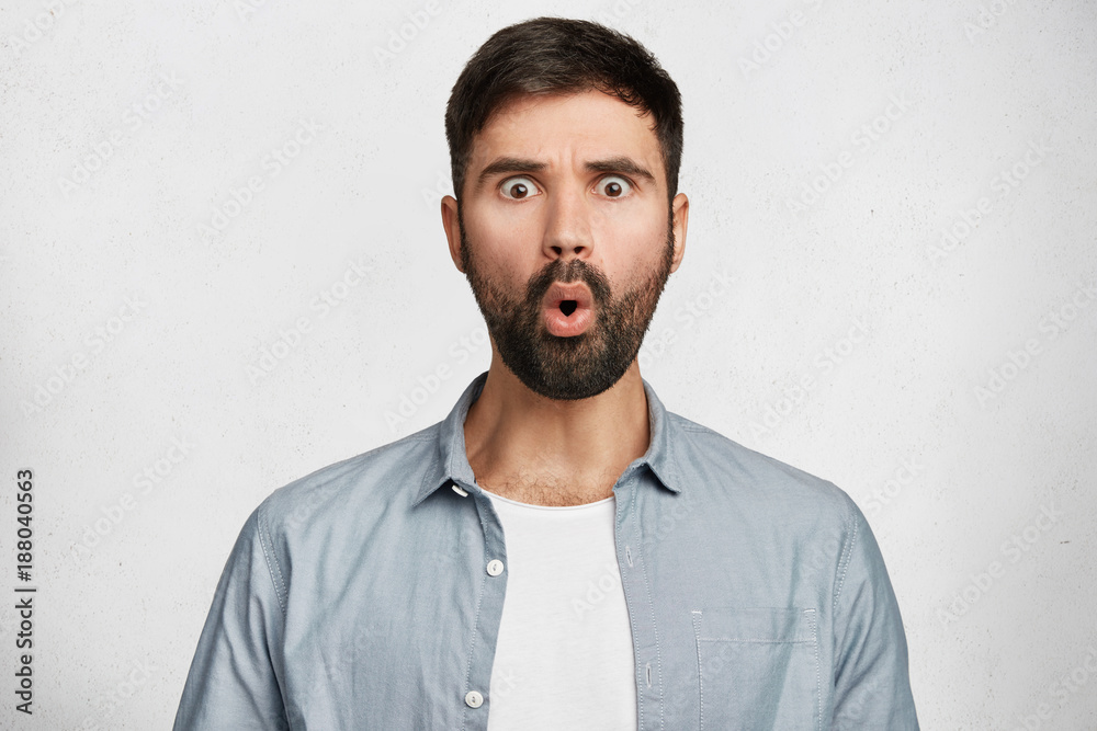Amazed bearded shocked man expresses great surprisment, dressed in fashionable clothes, being astonished to be promoted, poses against white background. Stunned unshaven male model in studio Amazed bearded shocked man expresses great surprisment, dressed in fashionable clothes, being astonished to be promoted, poses against white background. Stunned unshaven male model in studio