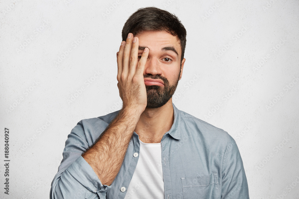Bearded male brunet covers face as shows his tiredness, curves lips, has no desire to work, dressed in denim shirt, isolated over white concrete background. Frustrated man with indignant look Bearded male brunet covers face as shows his tiredness, curves lips, has no desire to work, dressed in denim shirt, isolated over white concrete background. Frustrated man with indignant look
