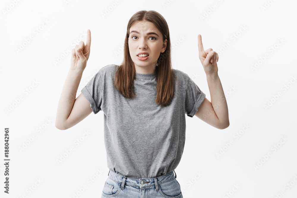 Portrait of young attractive dark-haired female student in fashionable outfit making funny face expression, pointing upside with index fingers.Copy space for advertisement. Portrait of young attractive dark-haired female student in fashionable outfit making funny face expression, pointing upside with index fingers.Copy space for advertisement.