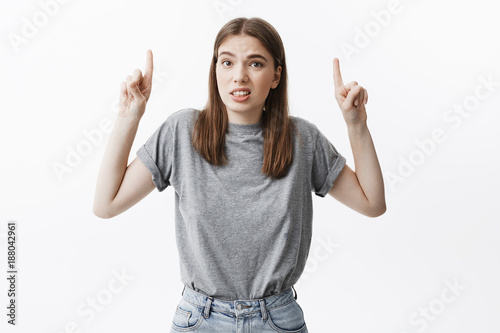 Portrait of young attractive dark-haired female student in fashionable outfit making funny face expression, pointing upside with index fingers.Copy space for advertisement.