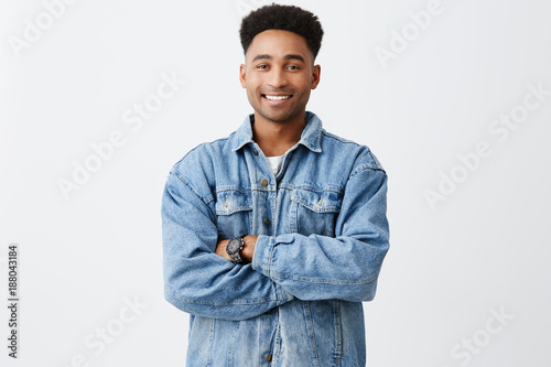 Isolated portrait of young funny dark-skinned man with arms crossed with afro hairstyle in casual white shirt under denim jacket with excited face expression