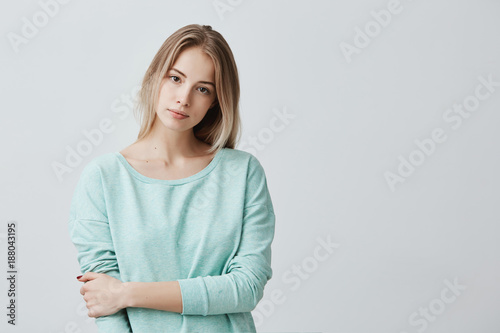 Portrait of young tender blonde european woman with healthy skin wearing light blue long-sleeved looking at camera with calm or pleasant expression. Caucasian female model posing indoors