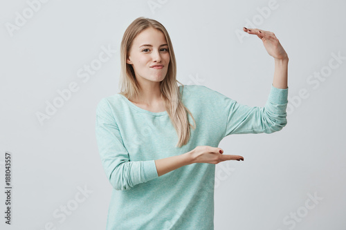 Young attractive woman in light blue sweater showing something big with hands, gesturing. Blonde female student demonstrating size of something against gray wall, expressing positive emotions.