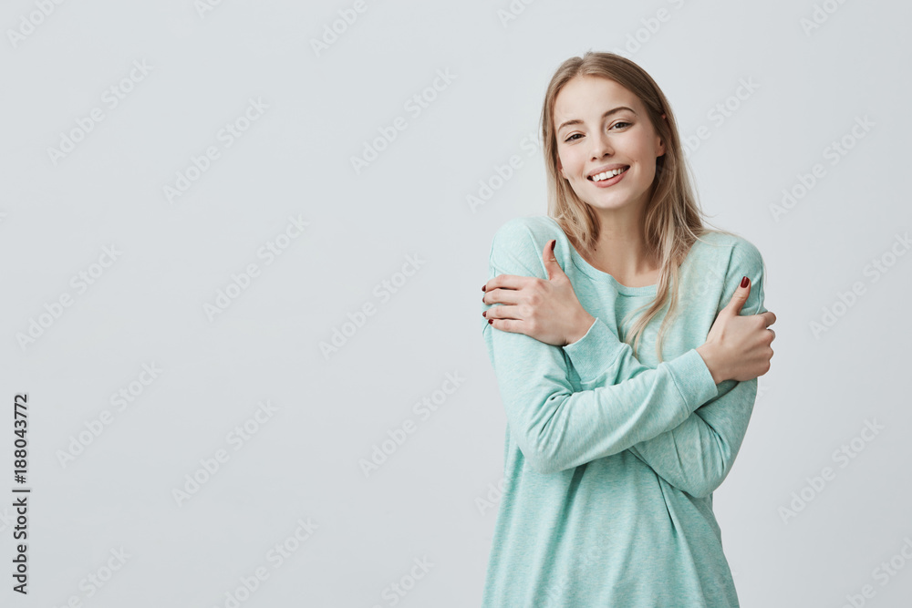 Waist up portrait of gorgeous positive young сaucasian female with charming smile and long blonde hair enjoying soft fabric of her new light blue top posing against gray wall Waist up portrait of gorgeous positive young сaucasian female with charming smile and long blonde hair enjoying soft fabric of her new light blue top posing against gray wall