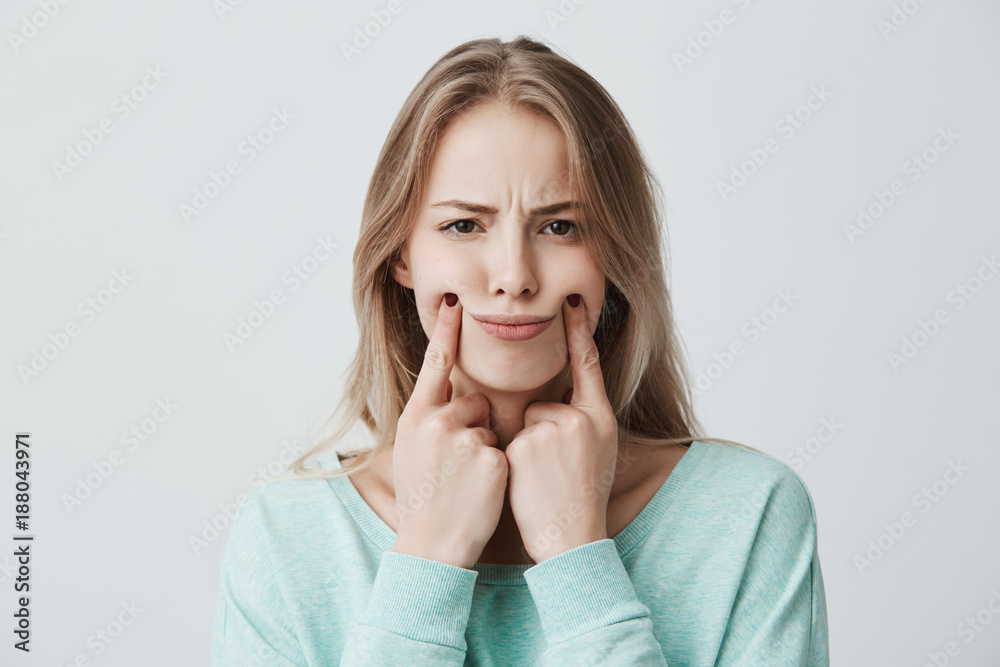Headshot of beautiful lovely young woman with dyed blonde hair, keeps hands on cheeks, has thoughtful and serious expression, dissatisfied and displeased, isolated against gray background Headshot of beautiful lovely young woman with dyed blonde hair, keeps hands on cheeks, has thoughtful and serious expression, dissatisfied and displeased, isolated against gray background