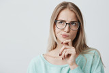 Dark-eyed blonde lovely young female with spectacles looks aside as has thoughtful expression and being concentraed on pleasant thoughts, dreams about something pleasant, has appealing appearance.