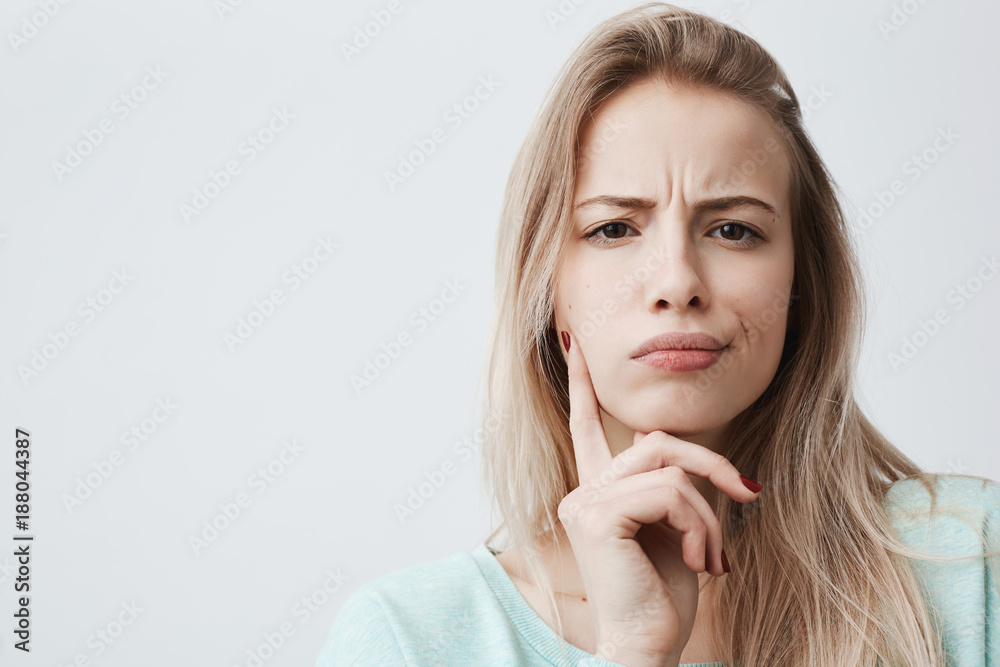 Horizontal portrait of displeased woman with blonde hair has indignant expression of face, frowns eyebrows, can`t understand something. Attractive puzzled dissatisfied female keeps hand on chin Horizontal portrait of displeased woman with blonde hair has indignant expression of face, frowns eyebrows, can`t understand something. Attractive puzzled dissatisfied female keeps hand on chin