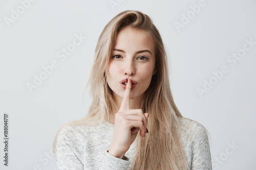 Shot of beautiful female with long blonde dyed hair wears casual long-sleeved t-shirt, shows hush sign, has serious expression, asks not to tell her secret anybody, hopes for loyality and silence. photo