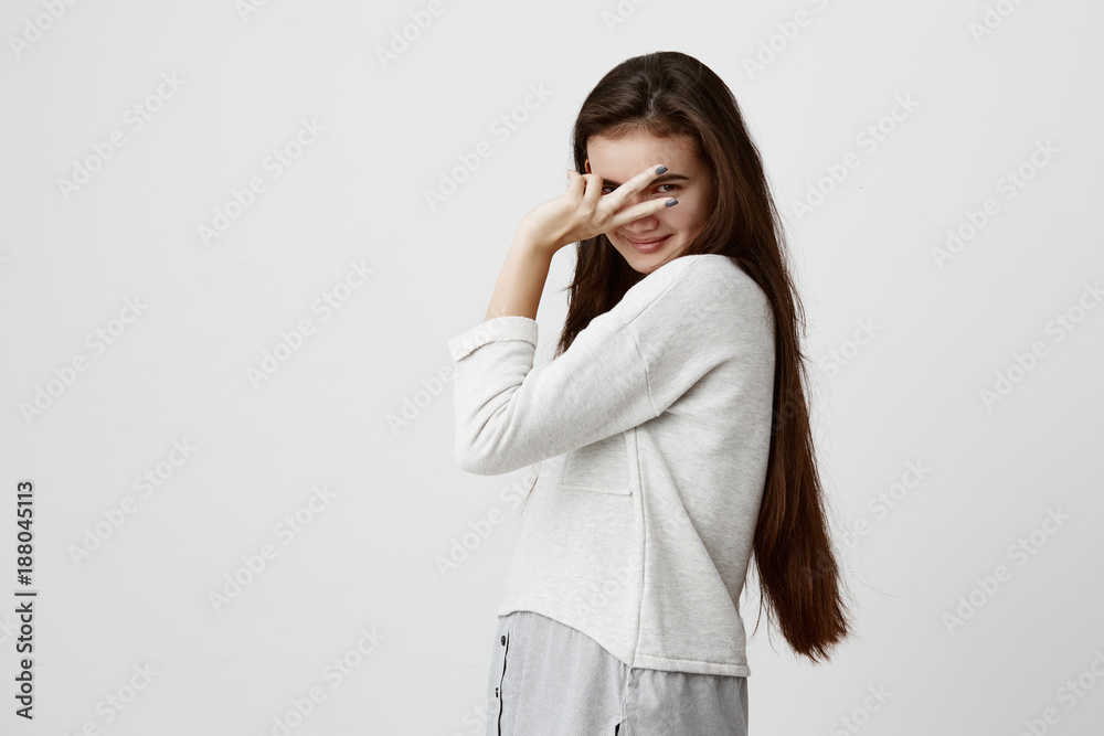 Portrait of beautiful female model with dark long hair, wears casual clothes, feels happy and relaxed indoors, showing V sign. Pretty girl smiles cheerfully, gestures, expresses positive emotions. Portrait of beautiful female model with dark long hair, wears casual clothes, feels happy and relaxed indoors, showing V sign. Pretty girl smiles cheerfully, gestures, expresses positive emotions.