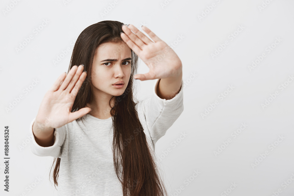 Body language. Stressed out angry beautiful teenage girl with dark long hair posing against gray studio wall, keeping hands in stop gesture, trying to defend herself as if saying: Stay away from me Body language. Stressed out angry beautiful teenage girl with dark long hair posing against gray studio wall, keeping hands in stop gesture, trying to defend herself as if saying: Stay away from me