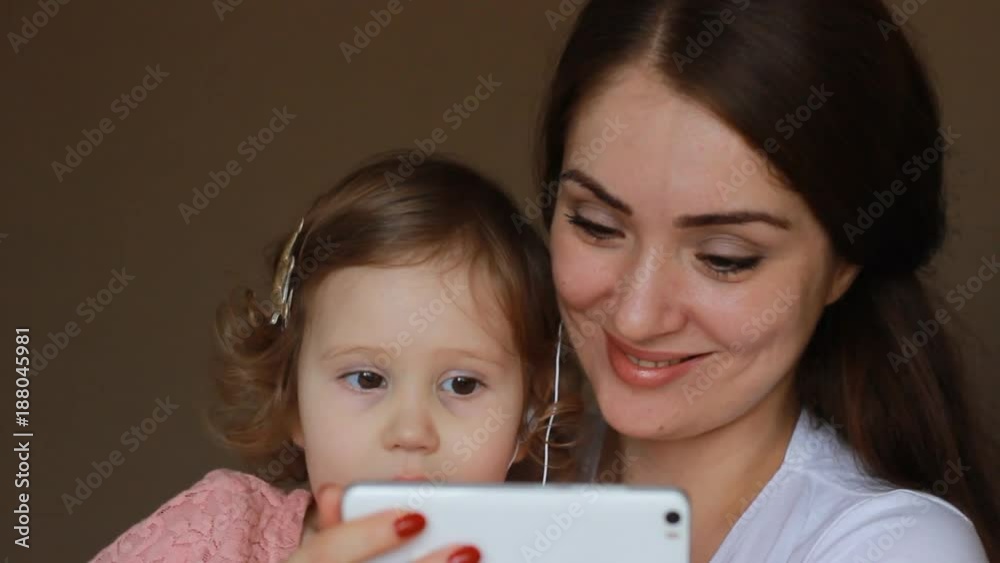 Mother and daughter in headphones look at the screen of the smartphone. Mom and child communicate by video link. The concept of the Internet, mobile phone and video chat Mother and daughter in headphones look at the screen of the smartphone. Mom and child communicate by video link. The concept of the Internet, mobile phone and video chat