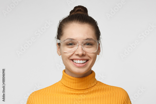 Close up shot of smiling and laughing attractive young woman in yellow polo neck sweater and transparent eyeglasses isolated on gray background photo