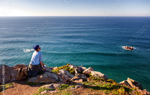 young beautiful woman sits on a stone on the background of Cape Cabo da Roca, Portugal, Coast of the Atlantic Ocean. Freedom, youth, travel