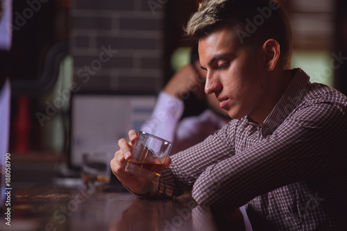 Side view - sad young hipster man looking at glass of whiskey and thinking about something sitting at the bar