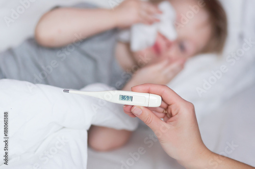 Close-up thermometer with fever heat 38. Mother measuring temperature of her ill kid at background. Sick child with high fever lying in bed at home. Mom Hand with thermometer close up