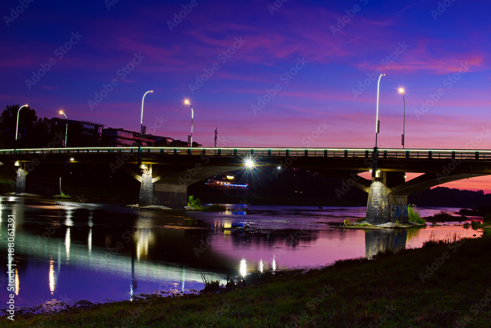 Big bridge through the river Uzh after sunset with illumination and reflection in the water. Beautiful cityscape of old european town Uzhgorod