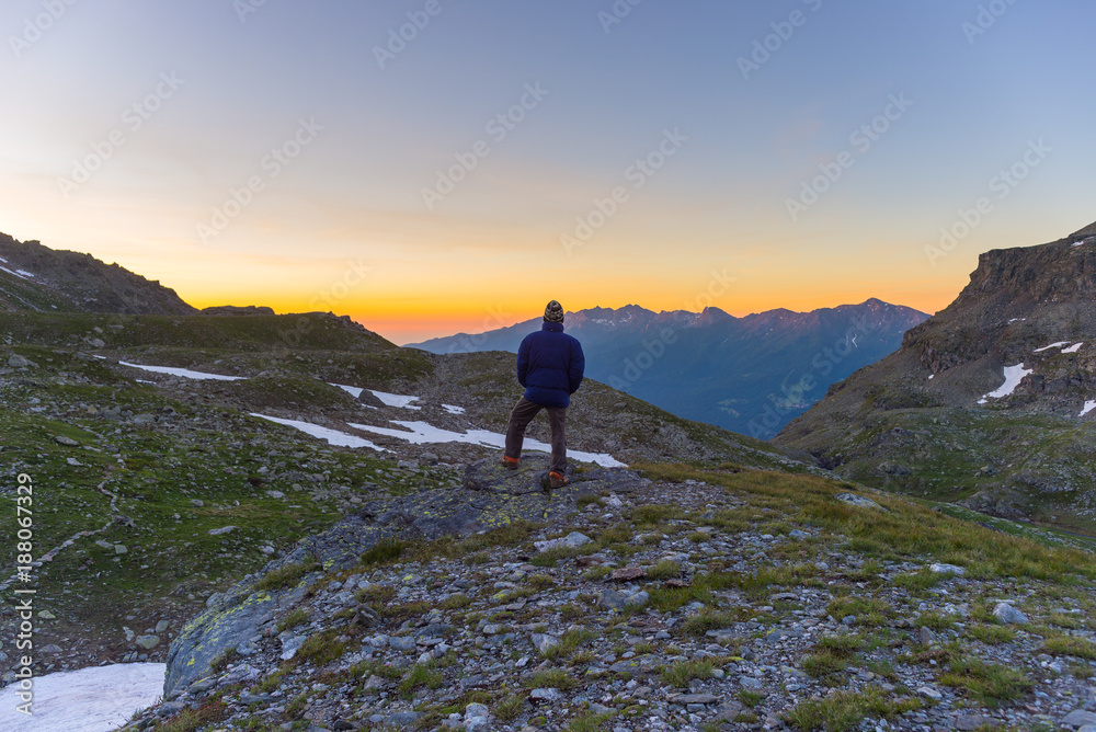 One person looking at colorful sunrise high up in the Alps. Wide angle view from above with glowing mountain peaks in the background. Summer adventure and exploration.