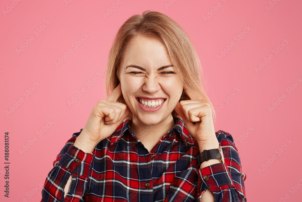 Studio shot of pretty woman clenches teeth and plugs ears with dissatisfaction as hears unpleasant sound or music, has appealing appearance, poses against pink background. Body language concept Studio shot of pretty woman clenches teeth and plugs ears with dissatisfaction as hears unpleasant sound or music, has appealing appearance, poses against pink background. Body language concept