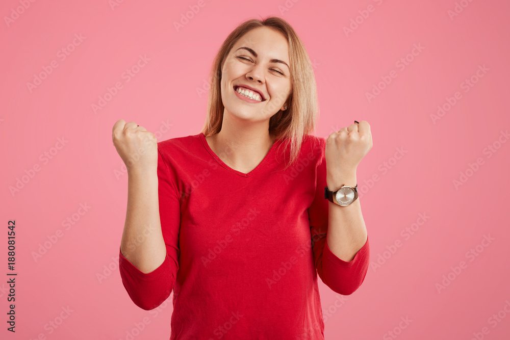 Glad female model clenches fists, dressed in red clothes, closes eyes with pleasure, dreams about something pleasant, isolated over pink background. Achievement, success and triumph concept. Glad female model clenches fists, dressed in red clothes, closes eyes with pleasure, dreams about something pleasant, isolated over pink background. Achievement, success and triumph concept.