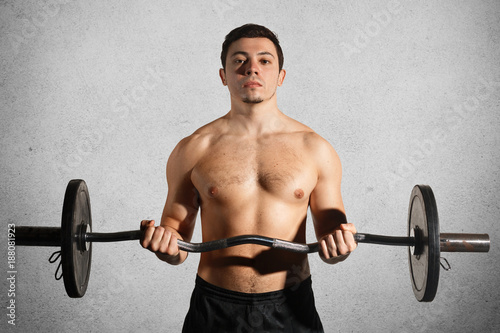 Sport, weightlifting, lifestyle and people concept. Confident sportsman with muscular body exercises in gym, lifts heavy weight, demostrates naked torso abs, trains biceps, isolated on grey wall