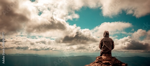 Tablou pe pânză Lonely man sitting on top of mountain cloudy sky