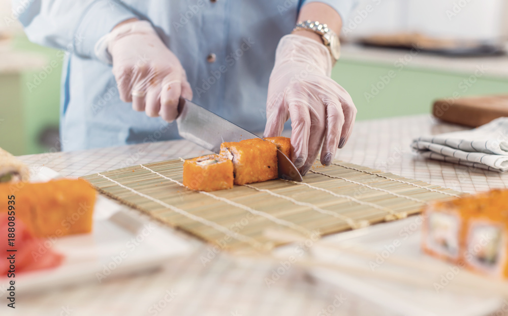Cropped image of woman in gloves cutting sushi rolls on bamboo mat and serving plates