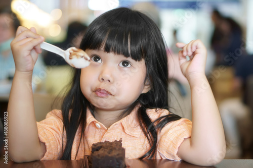Asian children cute or kid girl eating brownie chocolate cake for sweet dessert or snack with bored feeling and make face not delicious on wood table at lunch in restaurant or cafe