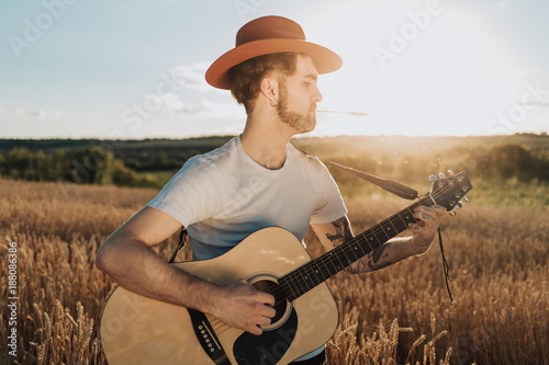 Young bearded man in hat with guitar enjoying music on a golden wheat field.