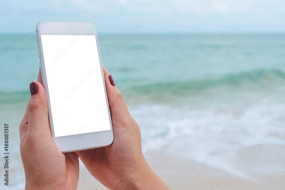 Mockup image of a hand holding and showing white mobile phone with blank black desktop screen in front of the sea and blue sky background