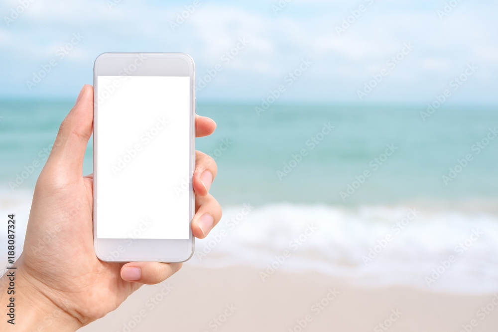 Mockup image of a hand holding and showing white mobile phone with blank black desktop screen in front of the sea and blue sky background