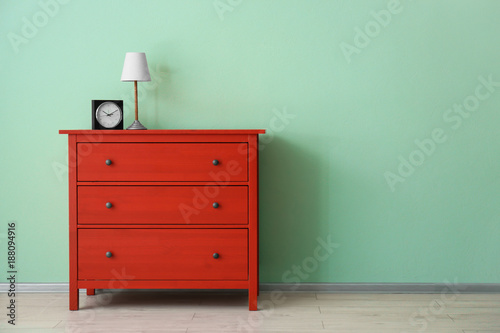 Red chest of drawers with clock and lamp near color wall © Africa Studio