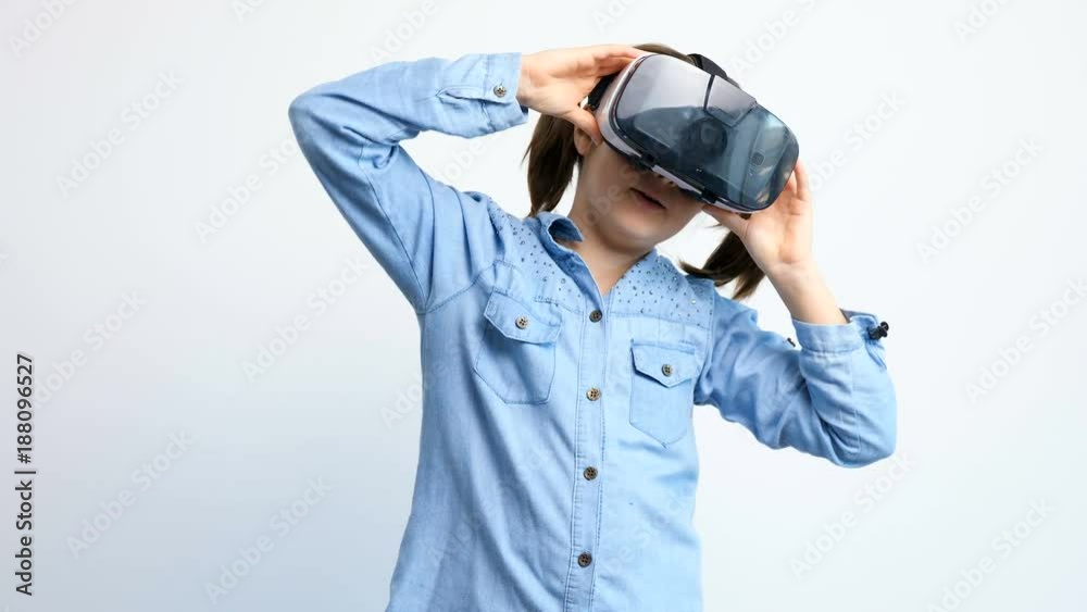 Cute young girl wearing VR virtual reality headset over a gray background playing a 360 degrees game and talking while doing it Cute young girl wearing VR virtual reality headset over a gray background playing a 360 degrees game and talking while doing it