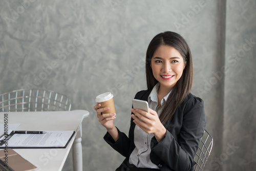 Asian business girl in her workstation at holding coffee cup and smartphone while smile