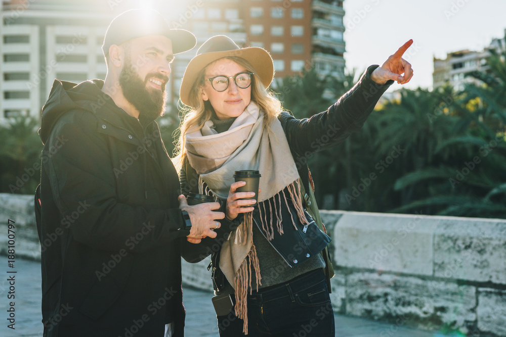 Sunny day. A loving couple of young tourists, friends are walking along the city street, drinking coffee. The girl is pointing at something. Vacation, travel, adventure, lifestyle. Backlight.