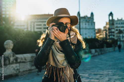 Sunny day, autumn. Young woman tourist, photographer, hipster girl dressed in hat and eyeglasses, stands on city street and takes photo. Vacation, travel, adventure, sightseeing. Blurred background.