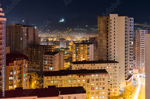 Top view of the night street of the sleeping area city of Batumi with high-rise buildings, light from the windows of residential houses, traffic of cars on the road. Urban life.