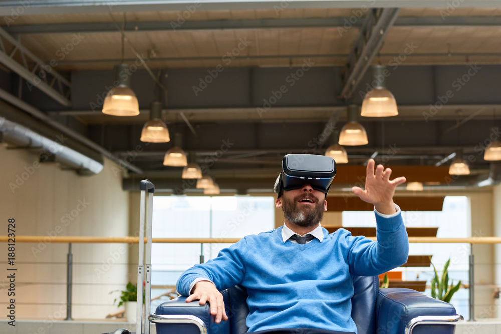 Portrait of mature bearded man wearing VR headset sitting in armchair looking amazed and excited reaching out with his hands, copy space