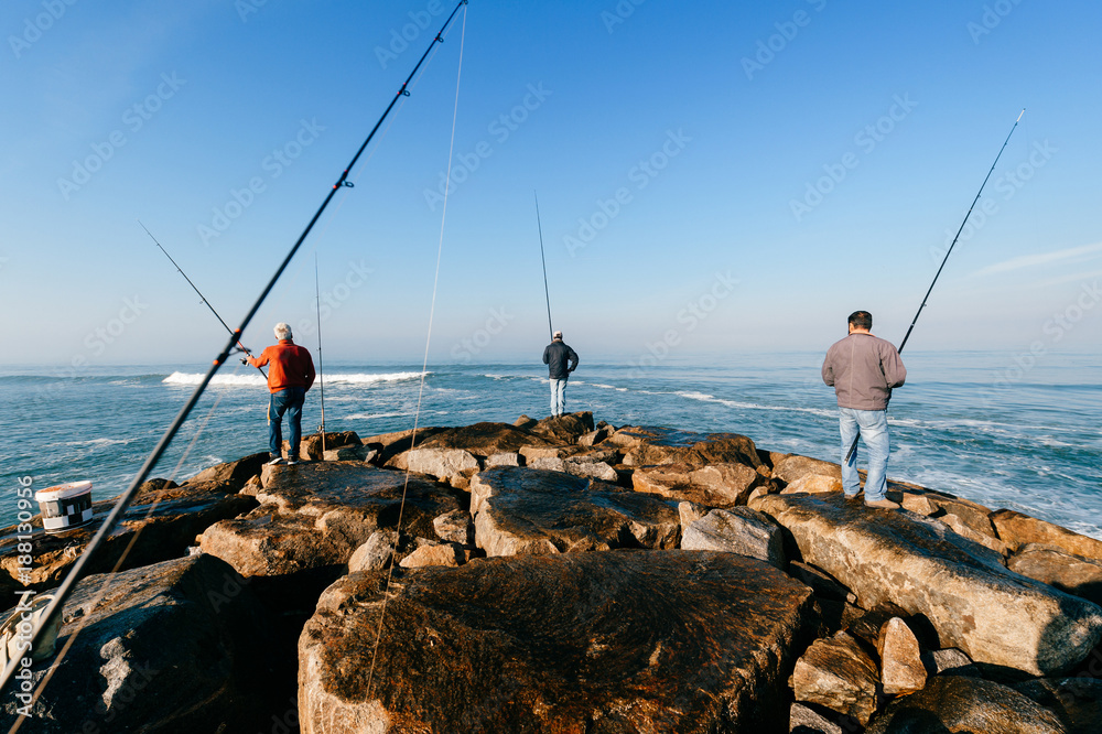 Morning at atlantic ocean in Portugal. Group of unrecognizable adult men  fishing. Unknown fisherman with fishing rod. Fishing gear. Rocky pier.  People at sea from behind. Sunrise at sea. Huge stones. Stock