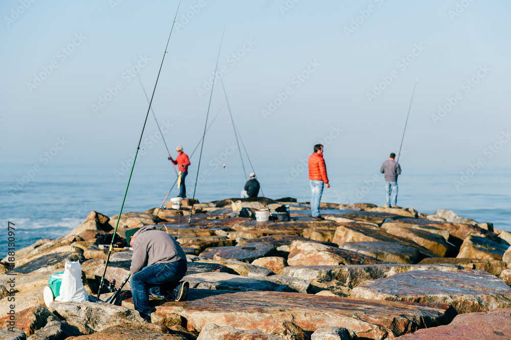 Morning at atlantic ocean in Portugal. Group of unrecognizable adult men  fishing. Unknown fisherman with fishing rod. Fishing gear. Rocky pier.  People at sea from behind. Sunrise at sea. Huge stones. Stock