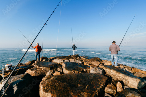 Morning at atlantic ocean in Portugal. Group of unrecognizable adult men fishing. Unknown fisherman with fishing rod. Fishing gear. Rocky pier. People at sea from behind. Sunrise at sea. Huge stones.