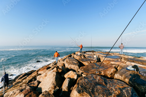 Morning at atlantic ocean in Portugal. Group of unrecognizable adult men fishing. Unknown fisherman with fishing rod. Fishing gear. Rocky pier. People at sea from behind. Sunrise at sea. Huge stones.