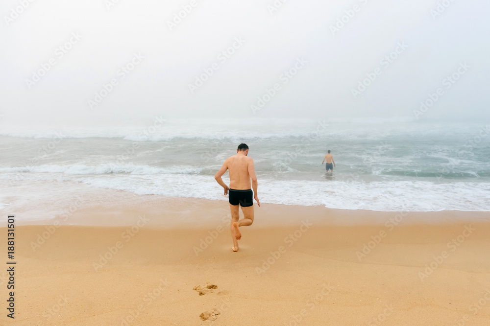 Topless man running from sandy beach in misty stormy ocean. Adult male enjoying swimming like child. Happy unrecognizable person. Water procedures. Healthcare and recreation. Crazy guy on vacation.