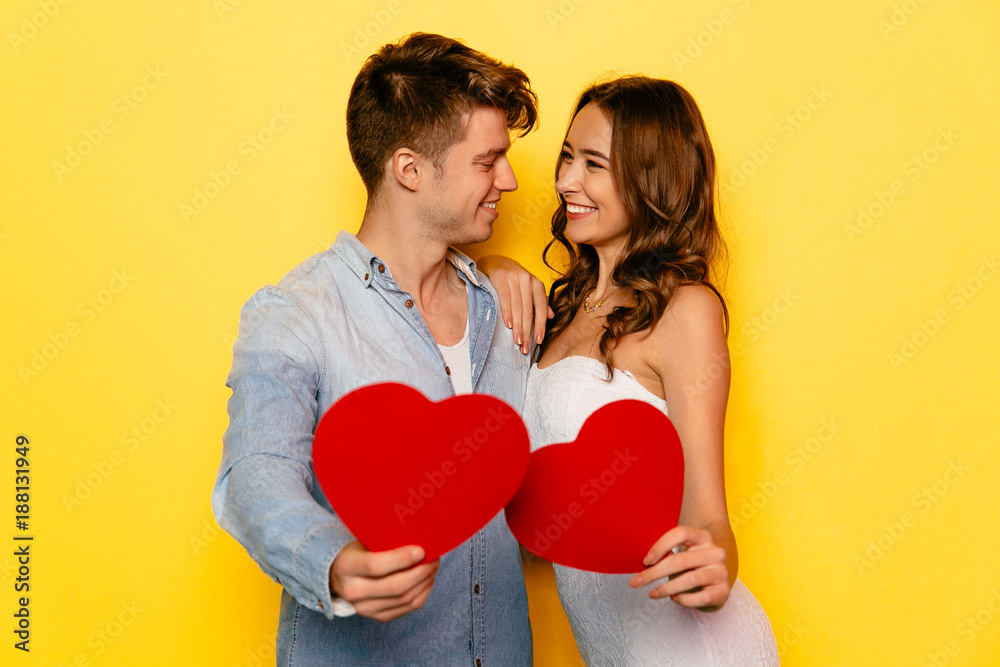 Cheerful smiling young couple celebrating St. Valentine's day, holding red hearts, looking to each other. Yellow background.