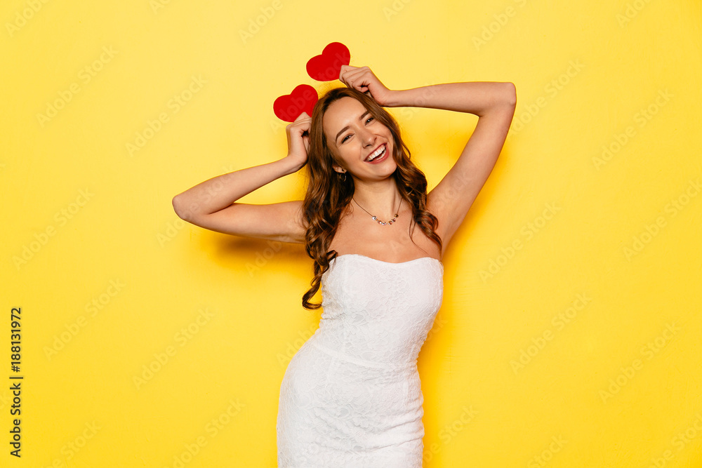 Beautiful funny girl has fun, while celebrating Valentine's day, holding red hearts above the head. Dressed in white dress, standing on yellow background.