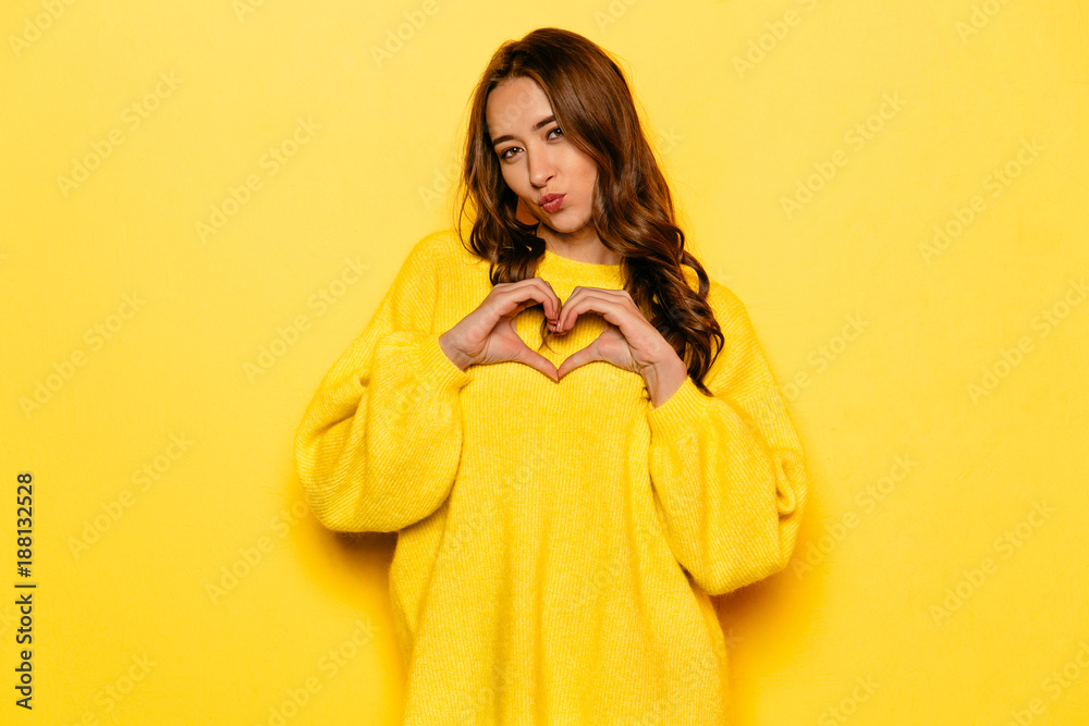 Charming young girl showing heart with two hands, love sign, giving a kiss, with curly hair. Dressed in yellow sweater. Charming young girl showing heart with two hands, love sign, giving a kiss, with curly hair. Dressed in yellow sweater.