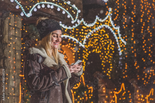 Young woman holding and looking at smart phone in front of house with christmas lights decoration