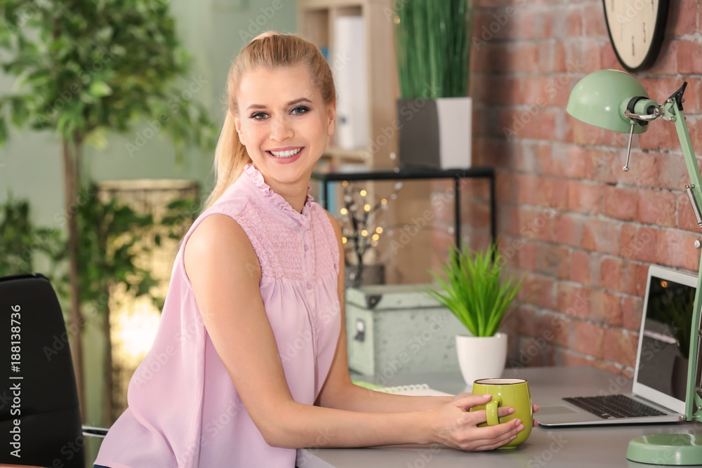 Young beautiful smiling woman with cup of coffee at table in office Young beautiful smiling woman with cup of coffee at table in office