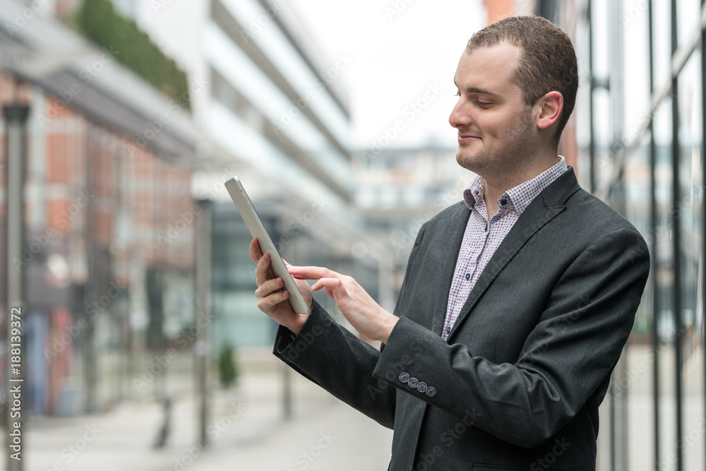 Young entrepreneur businessman smiling, checking and reading his email messages on his tablet, outdoors in the streets Young entrepreneur businessman smiling, checking and reading his email messages on his tablet, outdoors in the streets